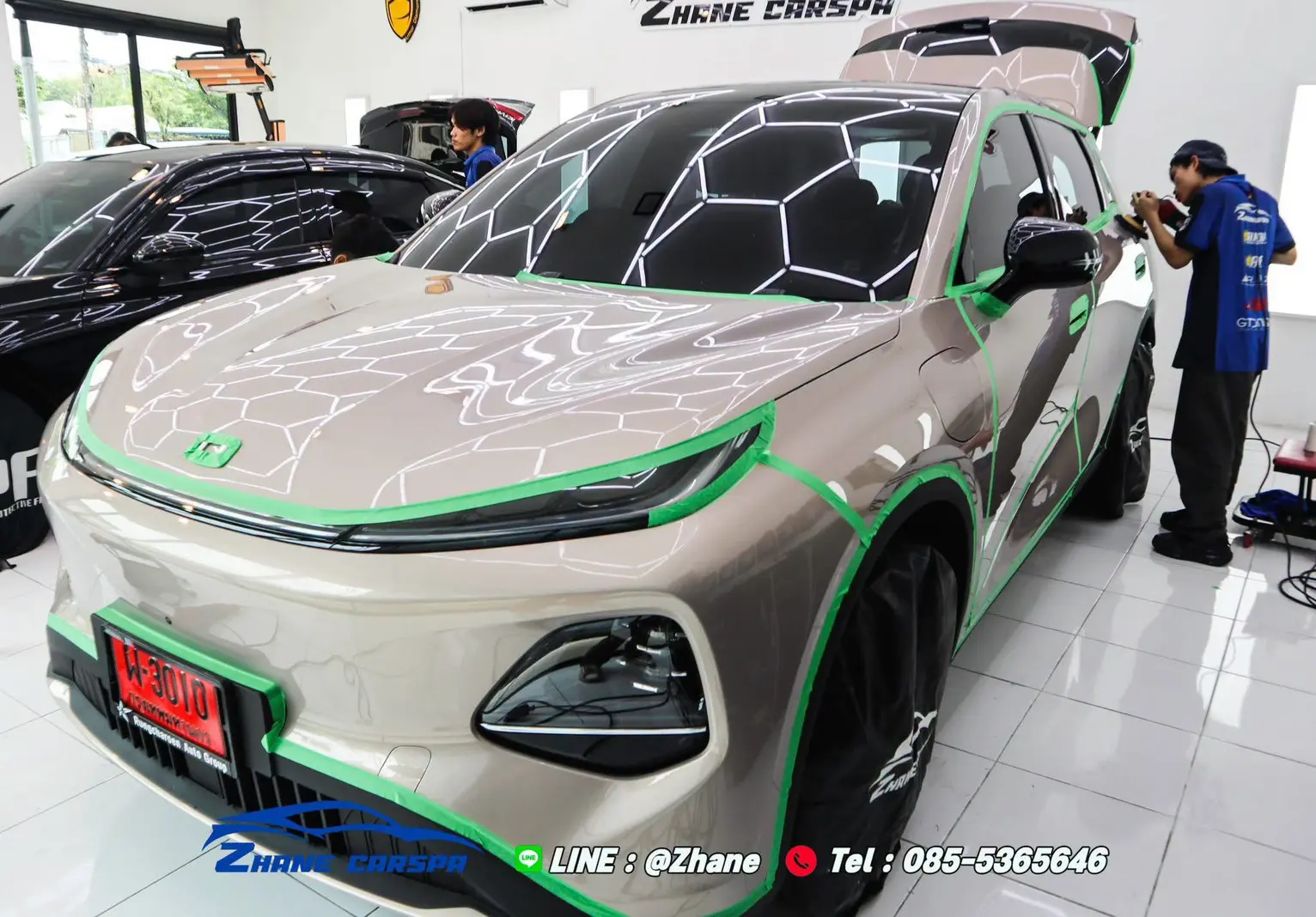 รถ SUV ไฟฟ้าสีแชมเปญติดเทปสีเขียวรอบไฟหน้า กันชน ซุ้มล้อ และกรอบป้ายทะเบียน ขณะช่างขัดสีด้านข้าง
