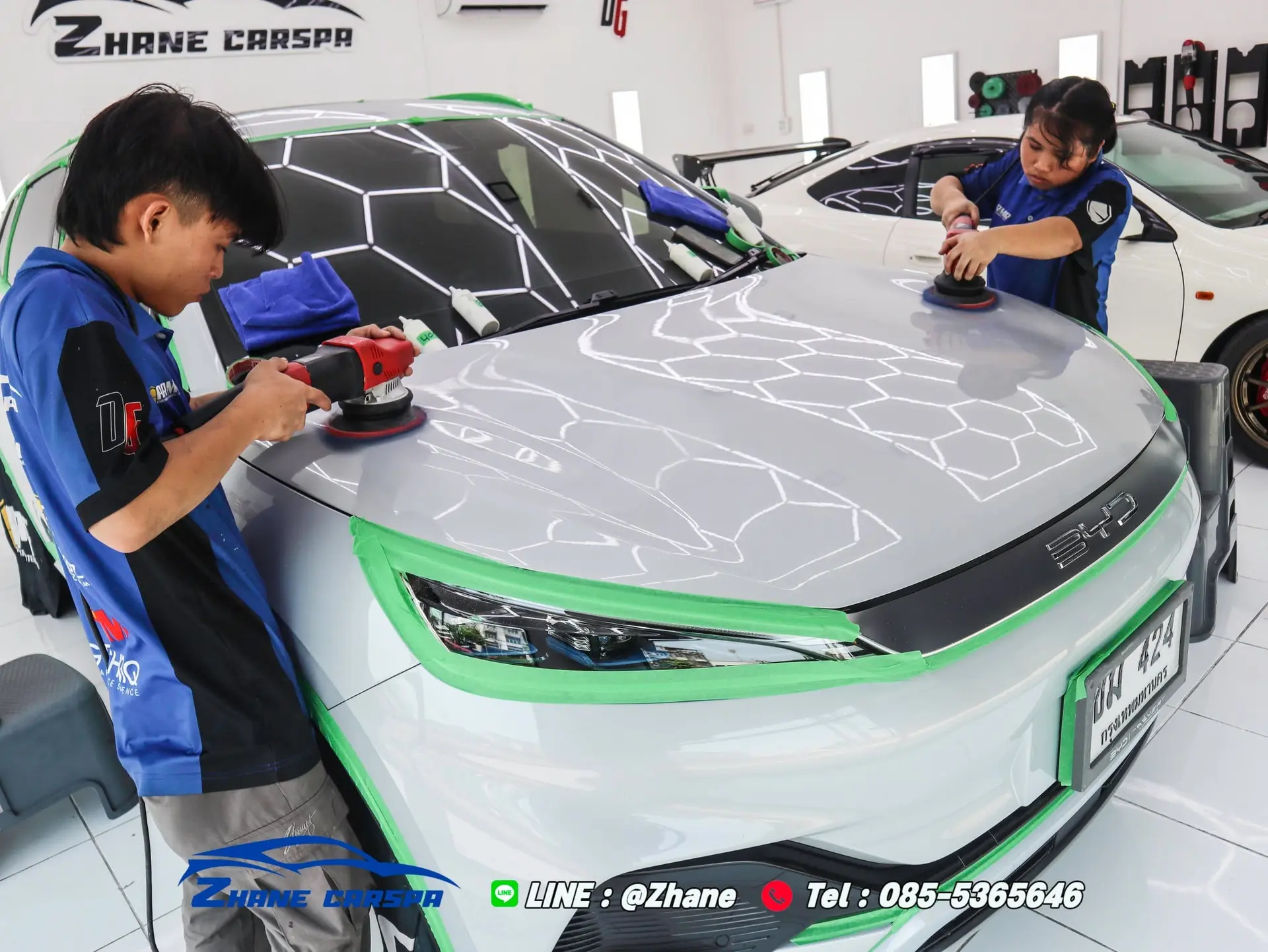 ช่าง Zhane Car Spa สองคนขัดเตรียมผิวฝากระโปรงรถ BYD Atto 3 สีขาวด้วยเครื่องขัด DA ขอบไฟหน้าติดเทปสีเขียว