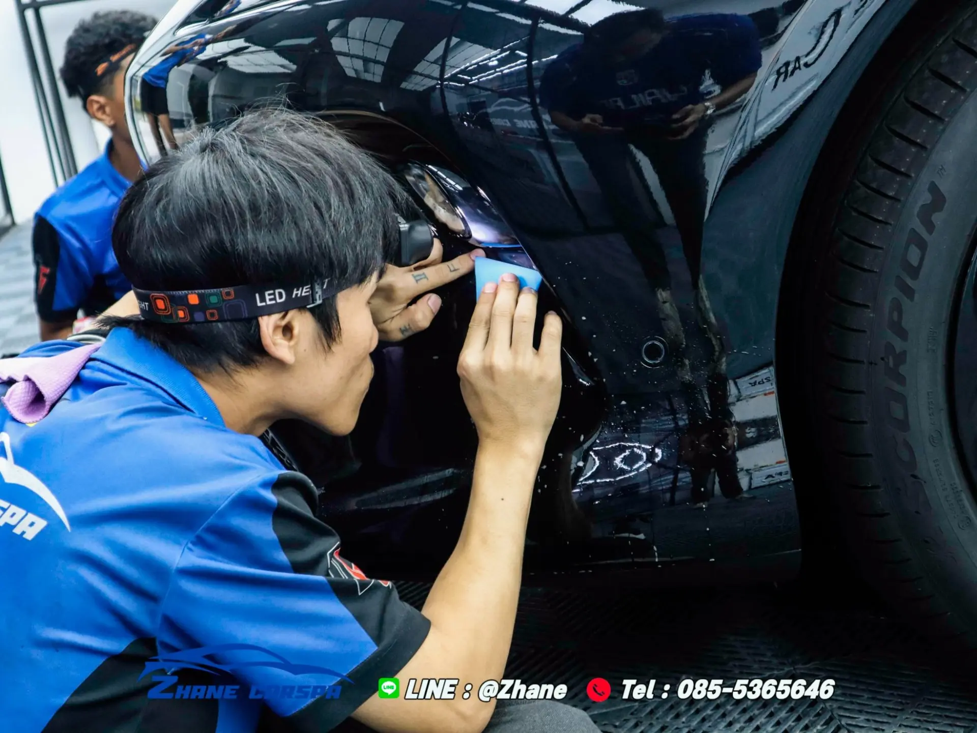 ช่าง Zhane Car Spa ใส่ไฟฉายคาดหัว ใช้เกรียงรีดพับขอบฟิล์มใสกันรอยเข้าใต้ขอบกันชนหน้ารถสีดำ